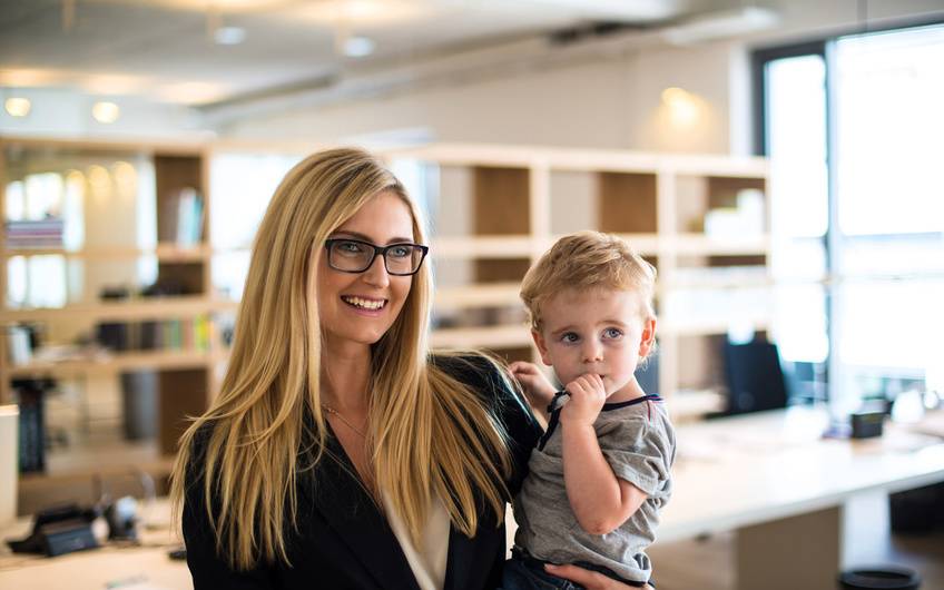 Eine blonde Frau in Business-Outfit trägt ihr kleines Kind im Arm durch die Büroumgebung.