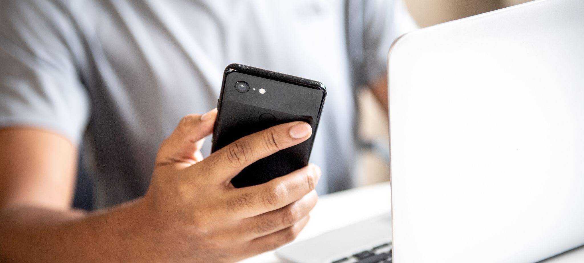Mann hält vor einem Laptop sein Smartphone in der Hand